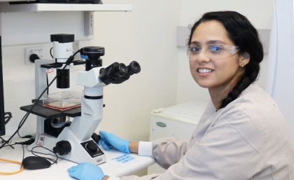 a woman with dark hair wearing safety glasses sits in front of a white and black microscope